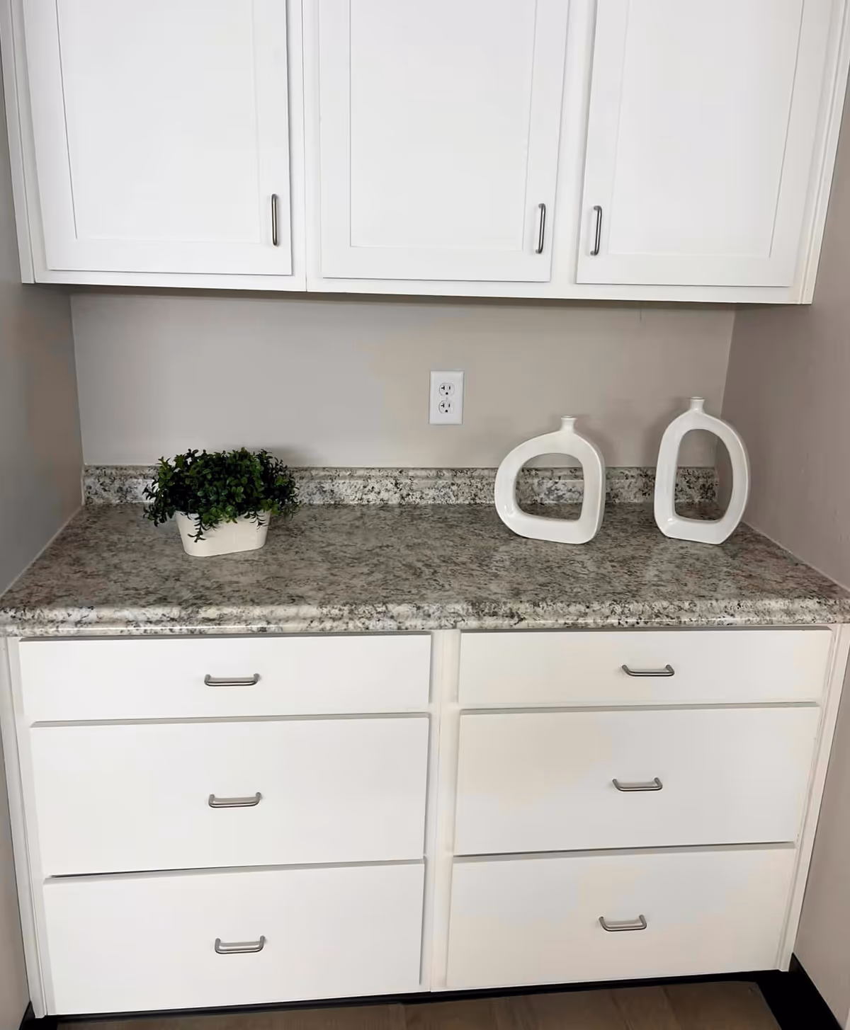 A kitchen countertop with a speckled granite surface, white cabinets above and below, a small green potted plant on the left, and two modern white decorative vases on the right.