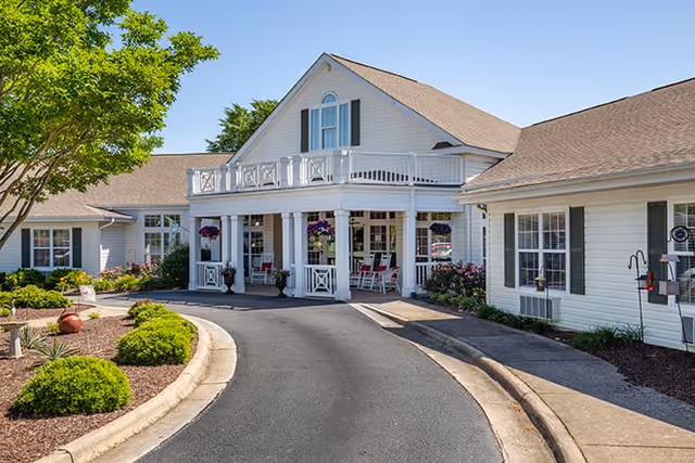 Front exterior view of a white senior living facility building with a covered entrance, a balcony above, hanging flower baskets, and a curved driveway surrounded by landscaped bushes and trees under a clear blue sky.