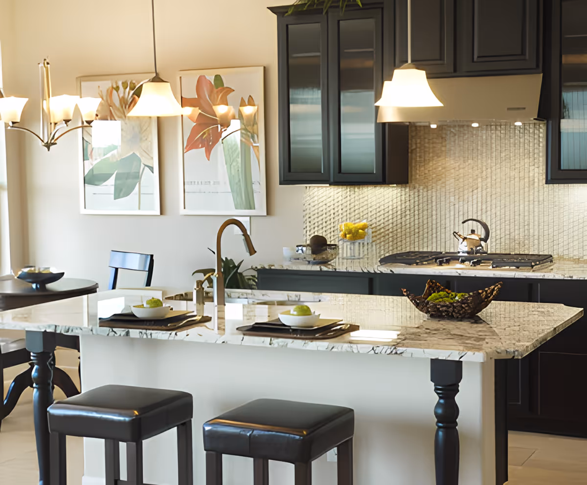 Modern kitchen with a marble island countertop featuring two black cushioned stools, a sink with a curved faucet, and decorative items. The background shows dark cabinetry, a stove with a kettle, a tiled backsplash, and two framed floral artworks on the wall. Pendant lights hang above the island and dining table.