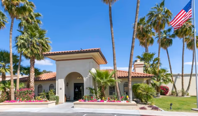 Front entrance of a single-story stucco building with a red tile roof, palm trees, flower beds, and an American flag.