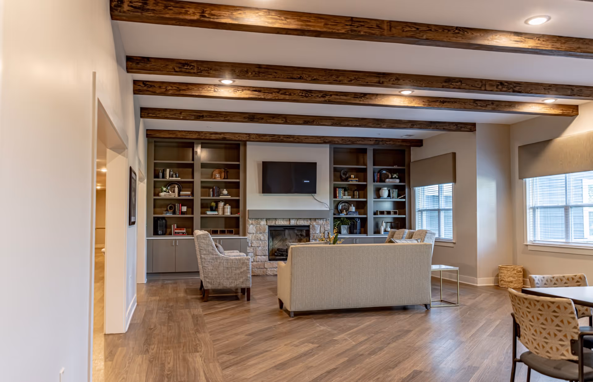 Cozy common lounge with sofas and armchairs facing a stone fireplace and built-in bookshelves beneath exposed wooden ceiling beams.