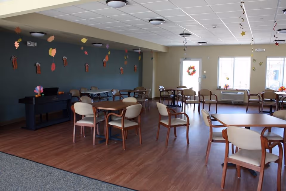 Communal dining/activity room with tables and chairs, a piano, and hanging fall decorations.