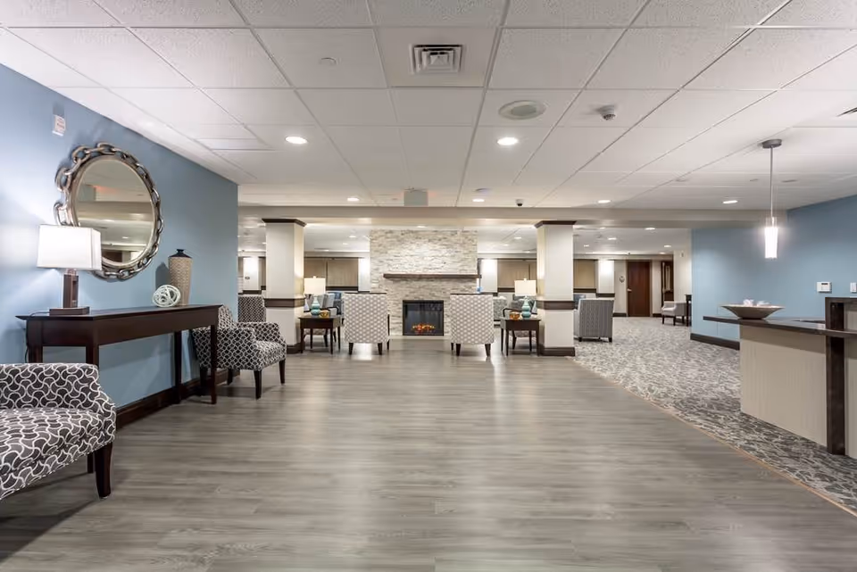 A spacious and well-lit senior living facility common area with light wood flooring and blue walls. The room features patterned armchairs, a console table with a lamp and decorative items, and a stone fireplace in the center background. The ceiling has recessed lighting and a hanging pendant light over a counter on the right side.