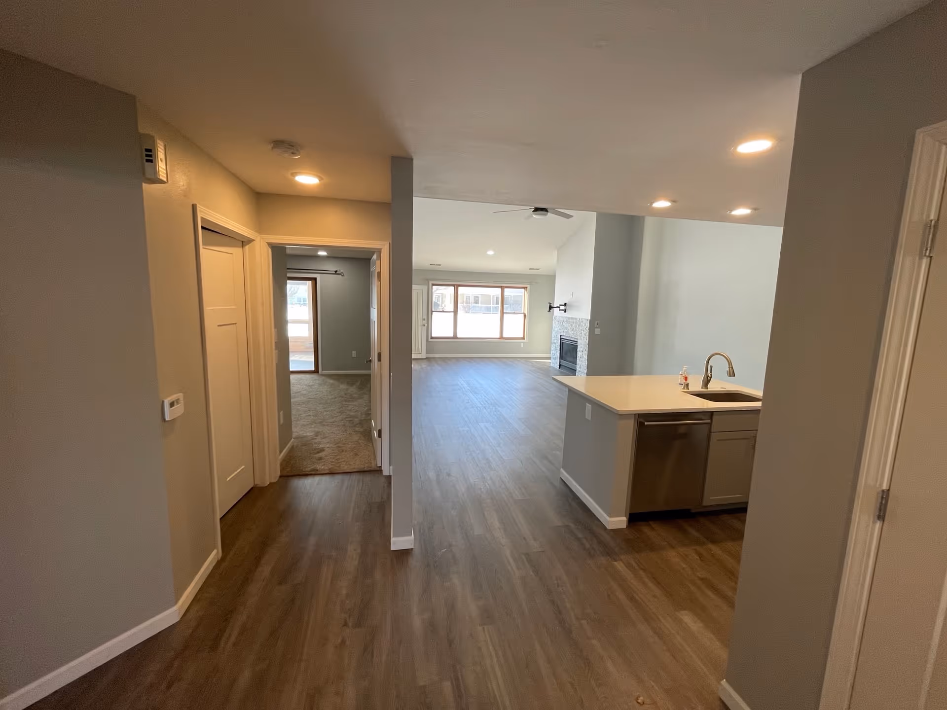 Interior view of a residential space showing a hallway leading to a carpeted bedroom on the left and an open living area with wood flooring, a kitchen island with a sink and dishwasher on the right, and a fireplace with a mounted TV bracket in the living room area.