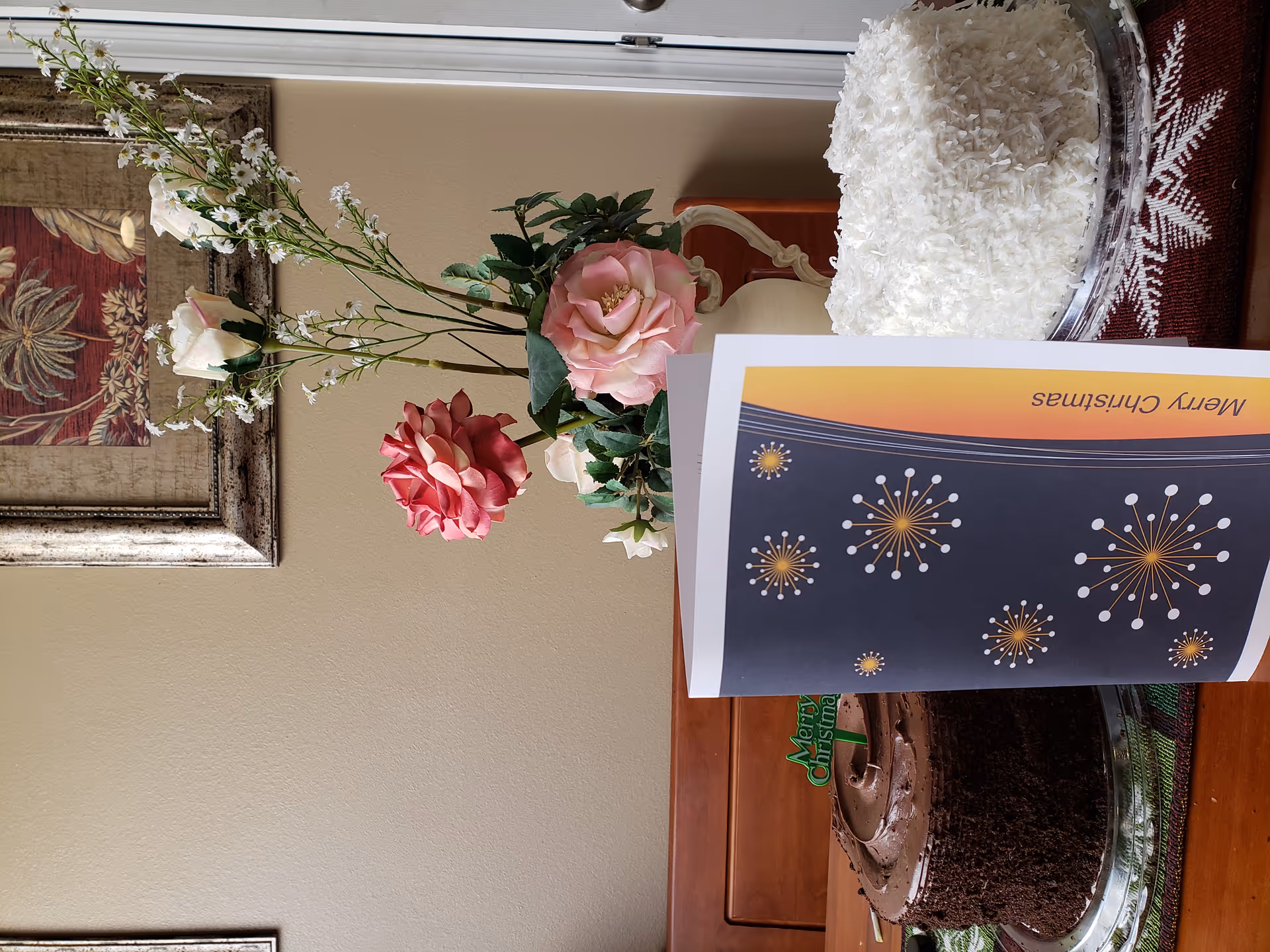 A table holding a vase of roses, two cakes (chocolate and coconut) and a 'Merry Christmas' card in a cozy interior with framed wall art.