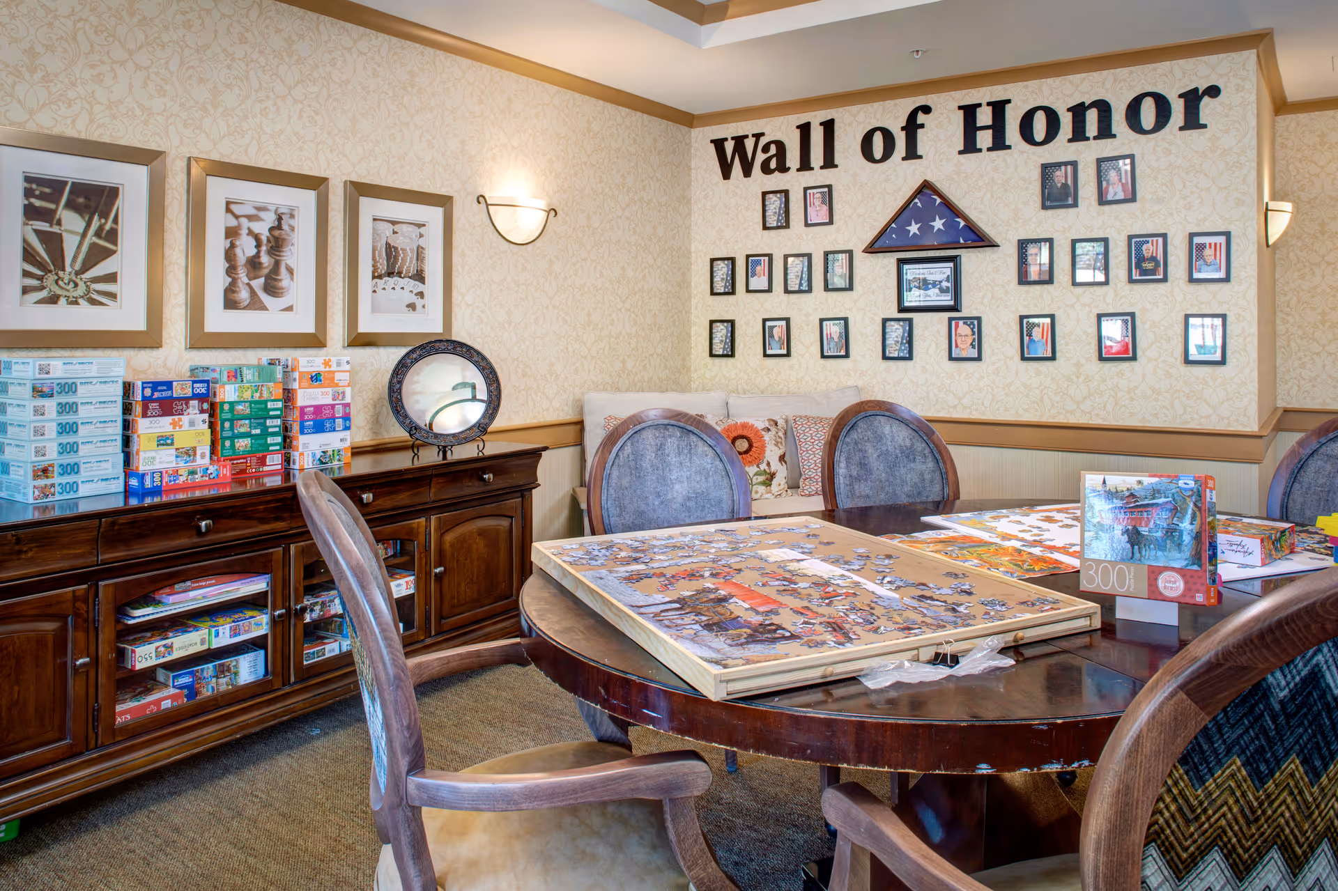 A cozy activity room with a round wooden table featuring an in-progress jigsaw puzzle. Surrounding the table are cushioned chairs. On the wall, there is a 'Wall of Honor' display with framed photos and a folded American flag. A wooden cabinet against the wall holds numerous puzzle boxes and board games. The room has warm lighting and patterned wallpaper.