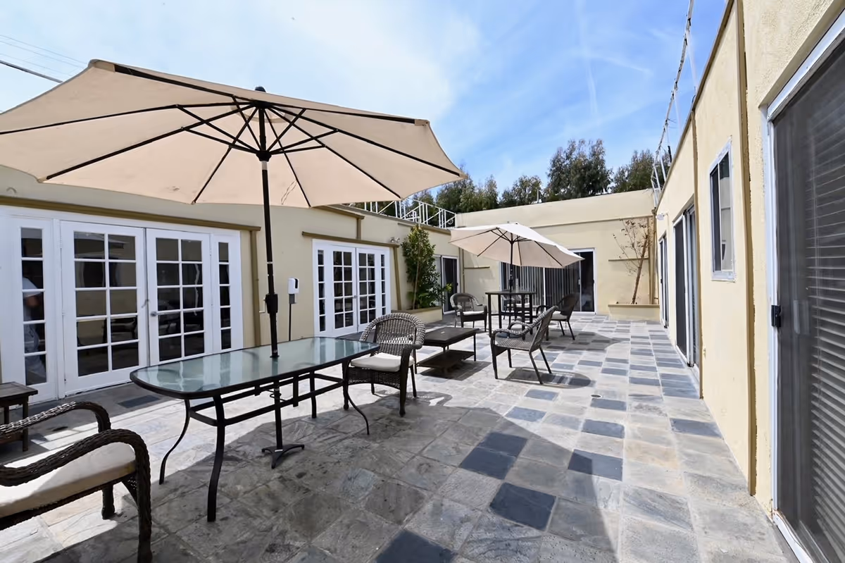 Outdoor patio area with stone tile flooring, featuring glass-top tables with large beige umbrellas and wicker chairs with cushions. The patio is surrounded by light yellow building walls with multiple white-framed glass doors and windows. Some greenery is visible along the walls under a partly cloudy blue sky.