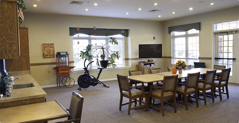 A bright communal dining/activity room with a long table and chairs, kitchenette, TV, exercise bike, and large windows.