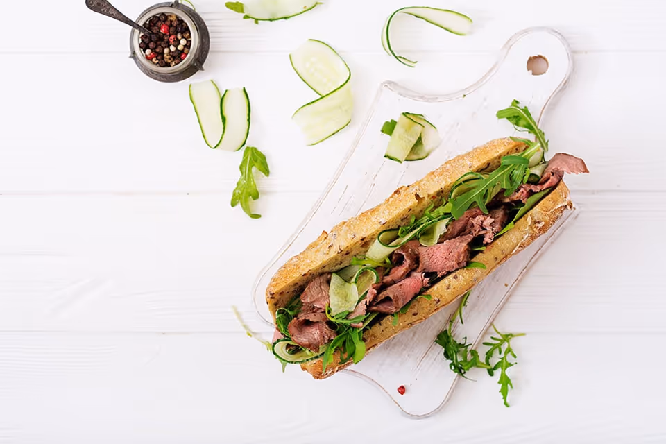 A sandwich on a white wooden cutting board filled with slices of roast beef, cucumber ribbons, and arugula, with a small container of mixed peppercorns and scattered cucumber slices and arugula leaves on a white wooden surface.