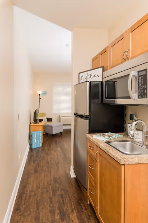 A narrow kitchen area with wooden cabinets, a countertop with a sink, a stainless steel refrigerator, and a microwave. Beyond the kitchen is a small living space with a floor lamp, a TV on a stand, a gray ottoman, and a window with blinds letting in natural light.