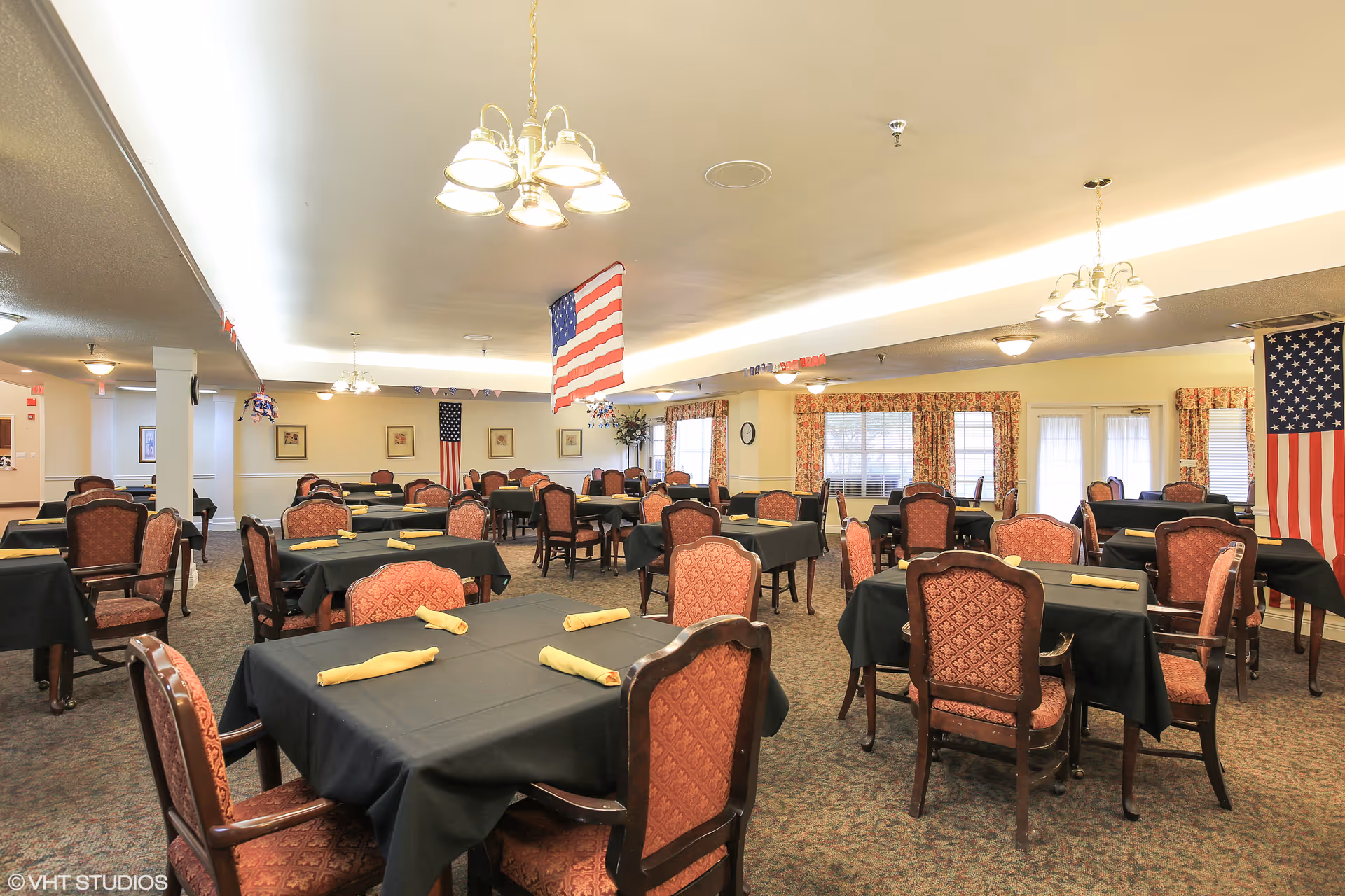 Spacious dining room with multiple tables covered in black tablecloths and yellow napkins, upholstered chairs, chandeliers, and American flags.