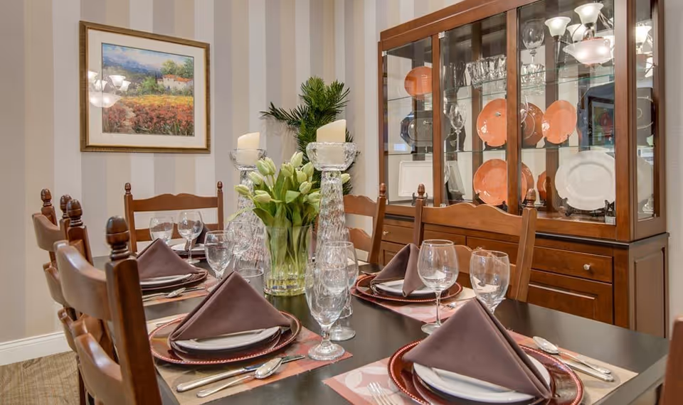 A formal dining room with a dark wooden table set for six people. Each place setting includes a folded brown napkin, a white plate, a red charger plate, and glassware. In the center of the table is a clear vase with white tulips and two tall glass candle holders with white candles. Behind the table is a wooden china cabinet displaying glassware and orange and white plates. The walls have beige and light brown vertical stripes, and a framed landscape painting hangs on the wall.