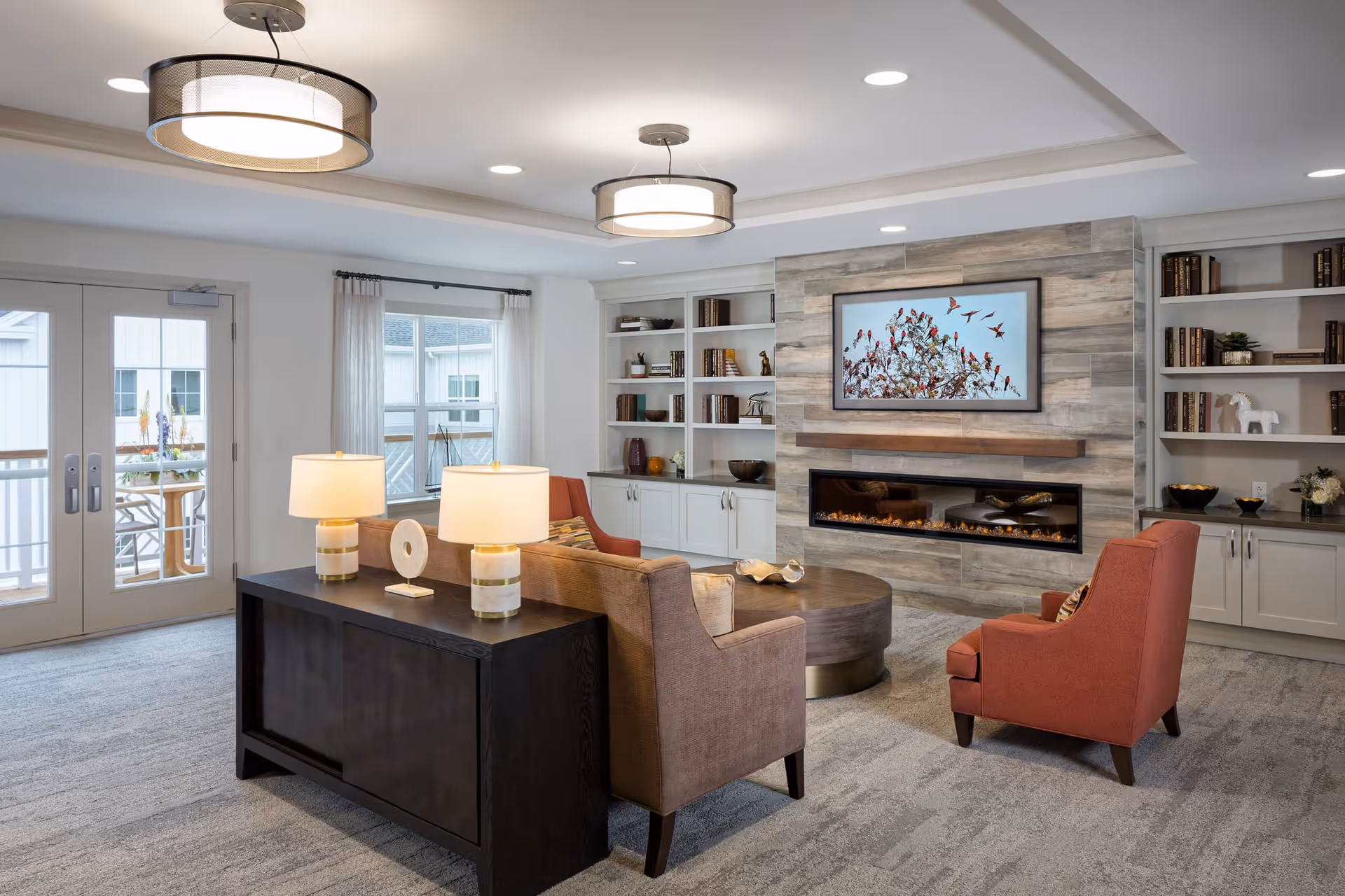 A cozy living room with a modern electric fireplace set in a wood-paneled wall, flanked by built-in white shelves filled with books and decorative items. The room features a brown sofa, an orange armchair, a round wooden coffee table, and a dark console table with two lamps. Large windows and glass doors allow natural light to enter, and two ceiling lights provide additional illumination.