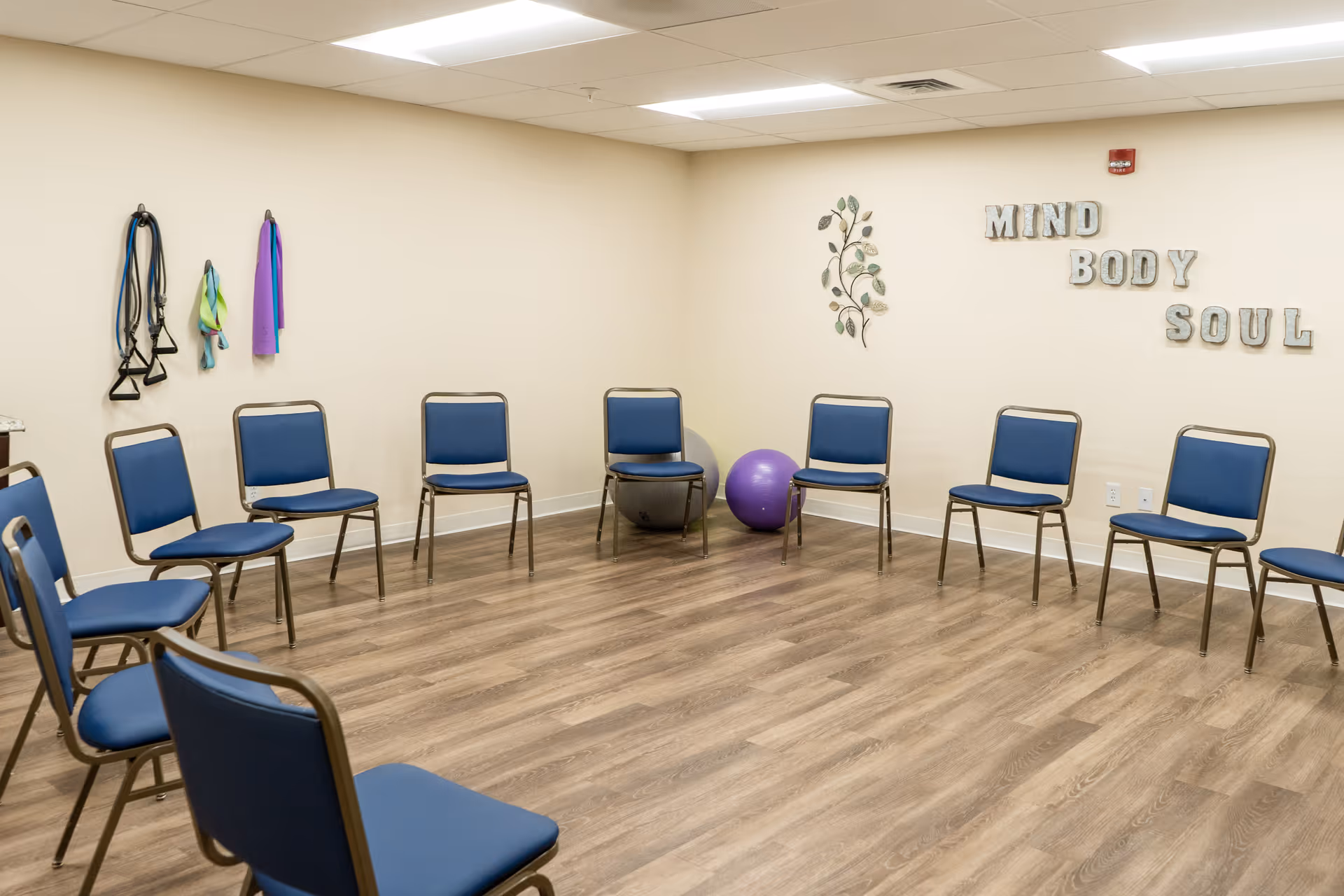 A room with chairs arranged in a circle on a wooden floor, exercise bands hanging on the wall, two exercise balls on the floor, and wall decor with the words 'MIND BODY SOUL'.