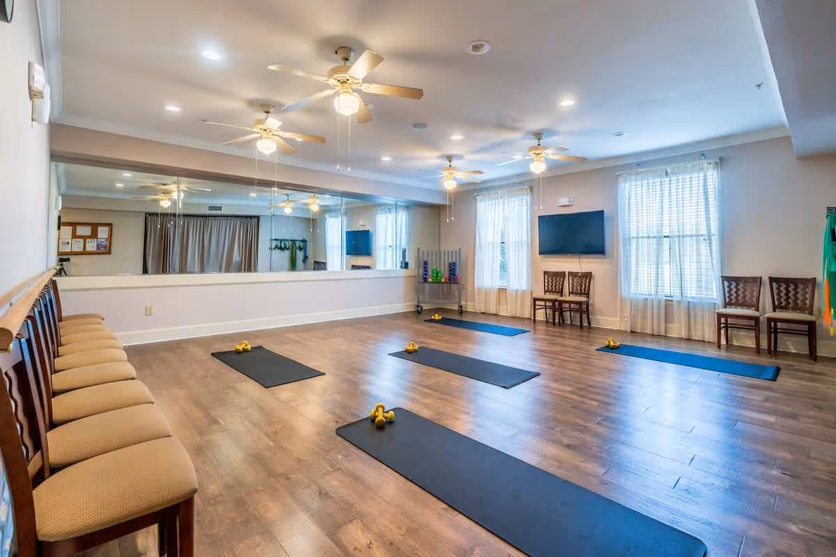 A spacious exercise room with wooden flooring, several ceiling fans, and large windows with sheer curtains. The room has a row of chairs along one wall and a large mirror covering another wall. Five black exercise mats are laid out on the floor, each with a pair of yellow dumbbells. There is a rack with colorful weights and a flat-screen TV mounted on the wall.