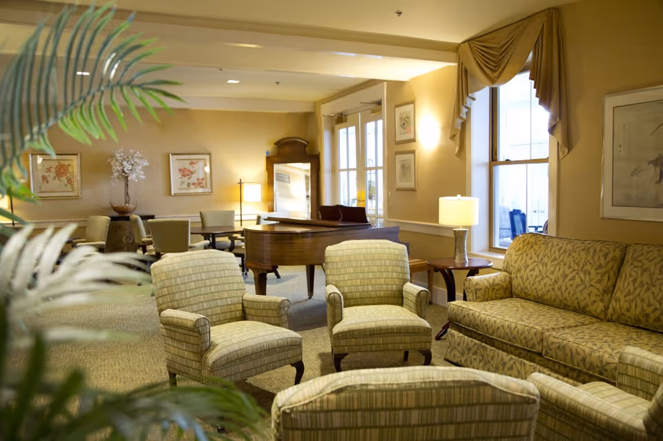 A cozy living room area with patterned armchairs and a matching sofa arranged around a wooden grand piano. The room is warmly lit with table lamps and wall sconces, decorated with framed floral artwork and a tall mirror. A large window with draped curtains lets in natural light, and a potted plant is partially visible in the foreground.