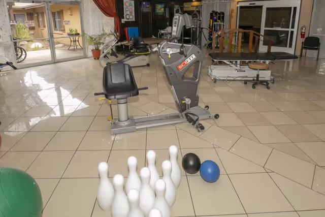 A tiled indoor rehabilitation room with exercise equipment, therapy tables, and a set of plastic bowling pins and balls on the floor.