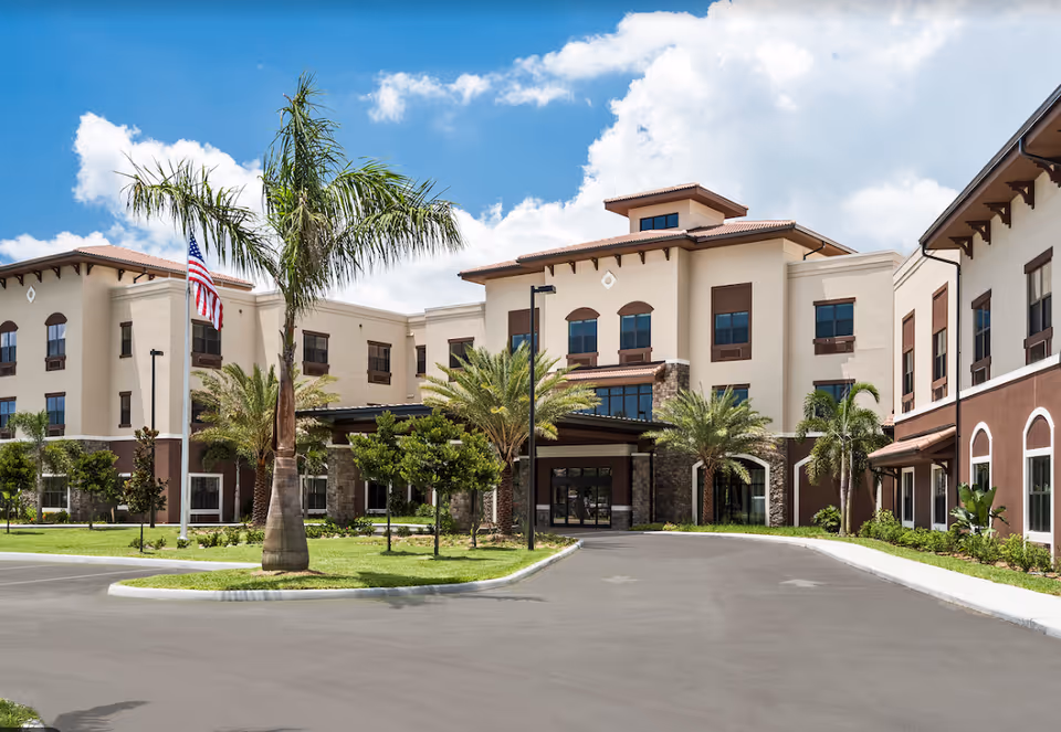 Front exterior of a three-story senior living building with a driveway, palm trees, and an American flag.
