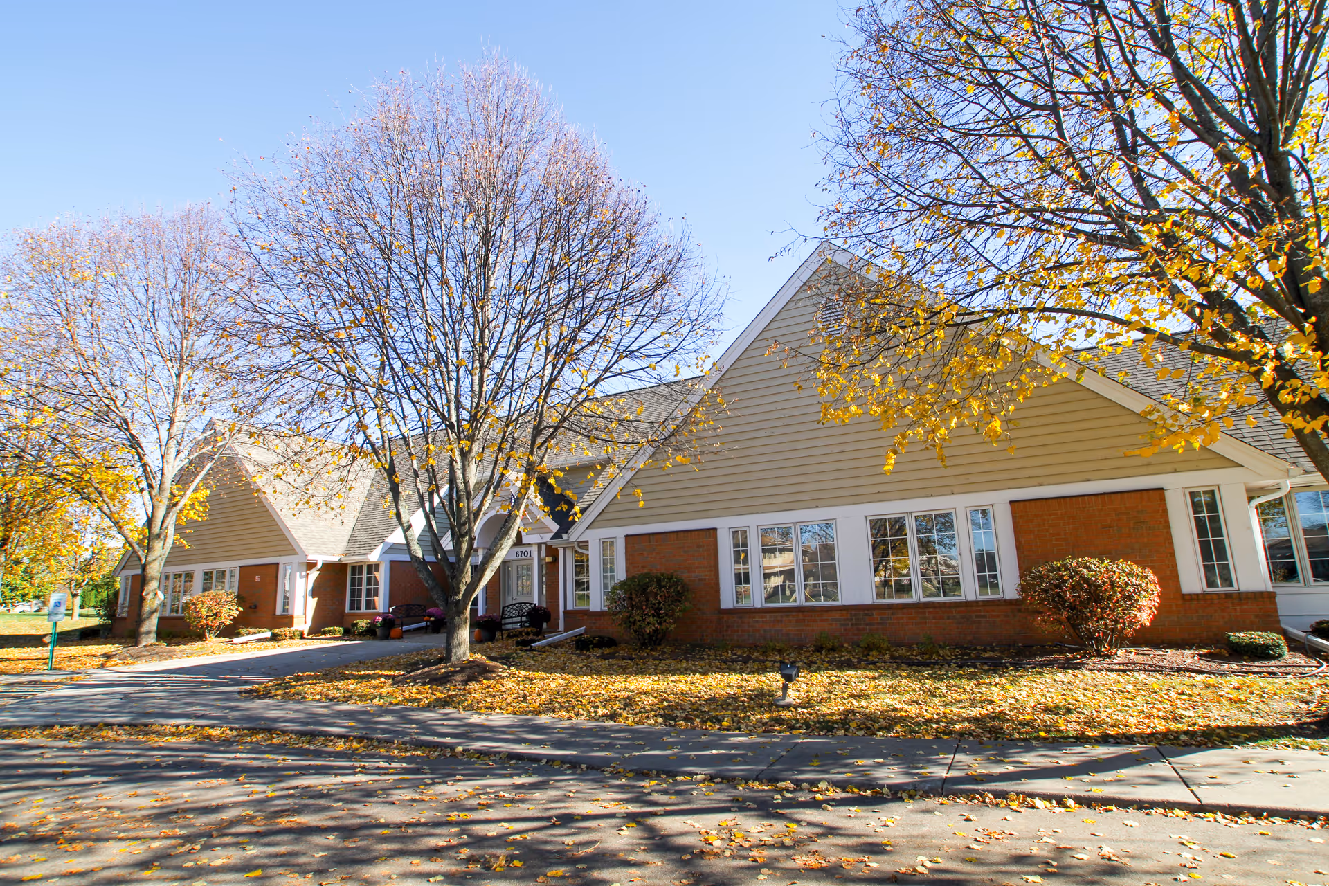 Single-story brick-and-siding senior living building front with bare autumn trees and fallen leaves on the lawn.