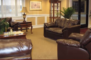 A cozy living room area with brown leather sofas and armchairs arranged around wooden side tables. The room has beige walls with white wainscoting, a framed picture on the wall, a table lamp, a tall glass display cabinet, and a potted plant near a glass door.