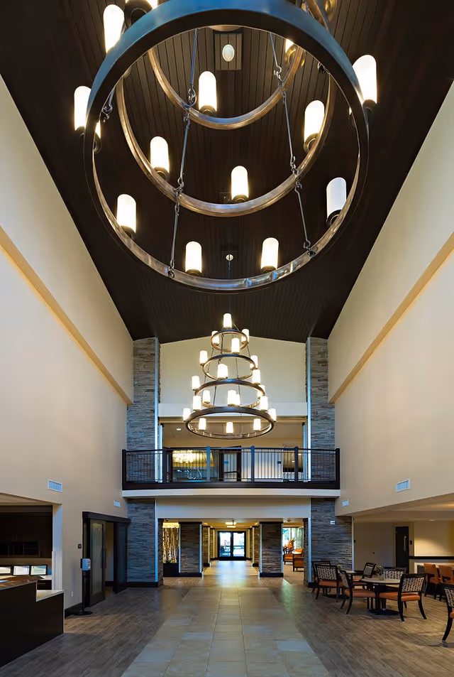 Spacious and well-lit interior lobby area with high ceilings featuring large circular chandeliers. The space includes a tiled floor, stone pillars, a balcony railing on the upper level, and seating areas with tables and chairs on the right side.