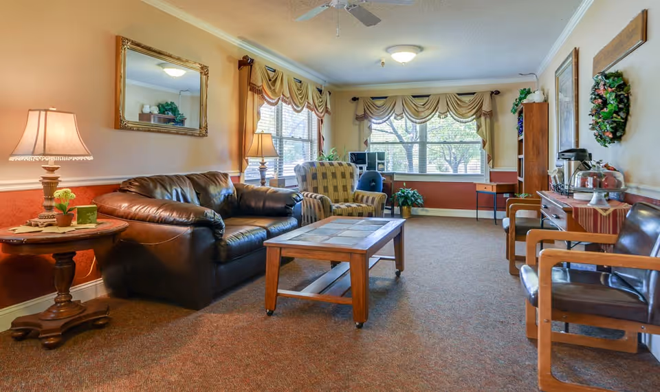 A cozy living room in a senior living facility with a brown leather couch, a patterned armchair, wooden coffee table, side table with a lamp, large windows with valances, and additional chairs along the wall. The room is warmly lit and decorated with plants and a wreath on the wall.