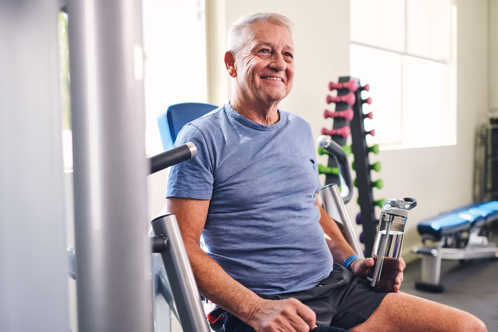 An elderly man wearing a blue t-shirt and black shorts is sitting on a gym machine in a fitness room, holding a water bottle and smiling. Behind him, there is a rack of colorful dumbbells and exercise benches near large windows letting in natural light.
