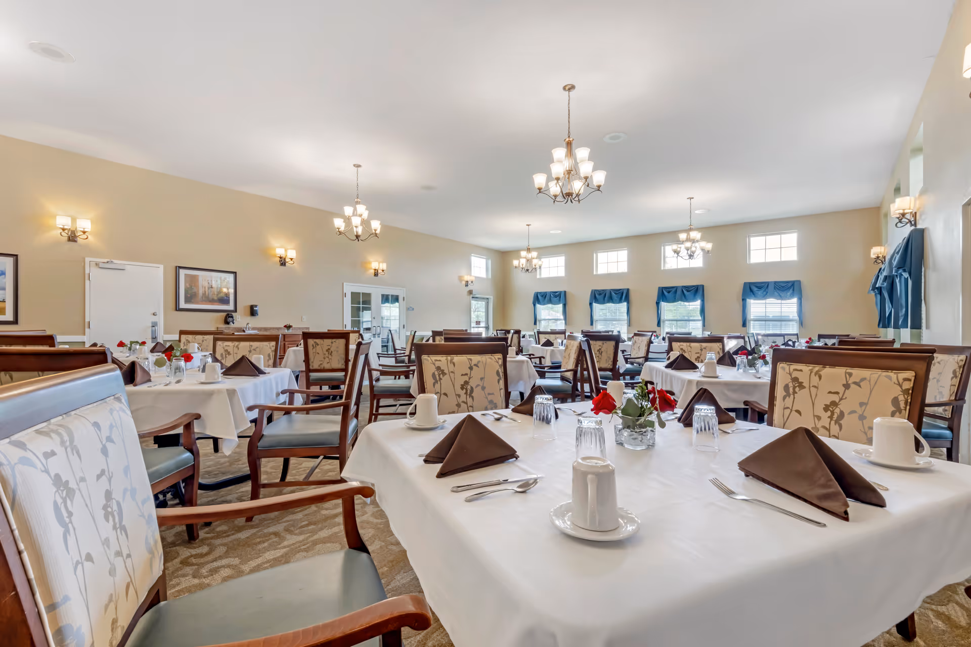 Spacious, well-lit dining room with multiple tables set with white linens, brown napkins, and floral centerpieces.