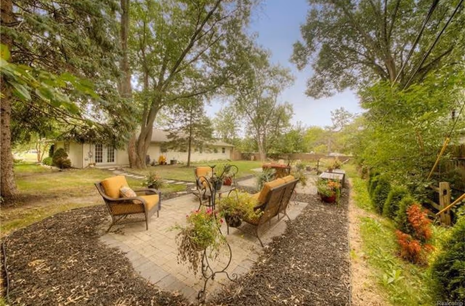 Outdoor patio area with cushioned wicker chairs and a small table surrounded by trees and plants, adjacent to a single-story building under a clear sky.