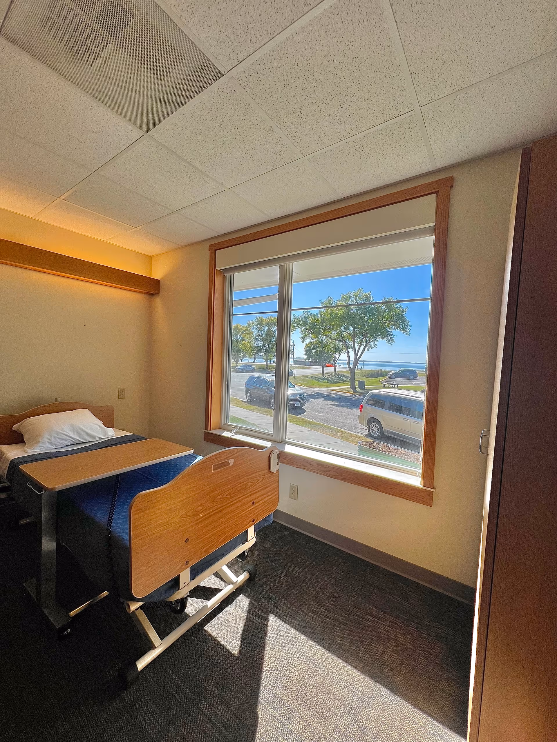 A small room with a single hospital-style bed with wooden headboard and footboard, a pillow, and a blue blanket. There is a wooden overbed table positioned over the bed. The room has a large window with a view of parked cars, trees, and a clear blue sky outside. The walls are light-colored, and the ceiling has white acoustic tiles.