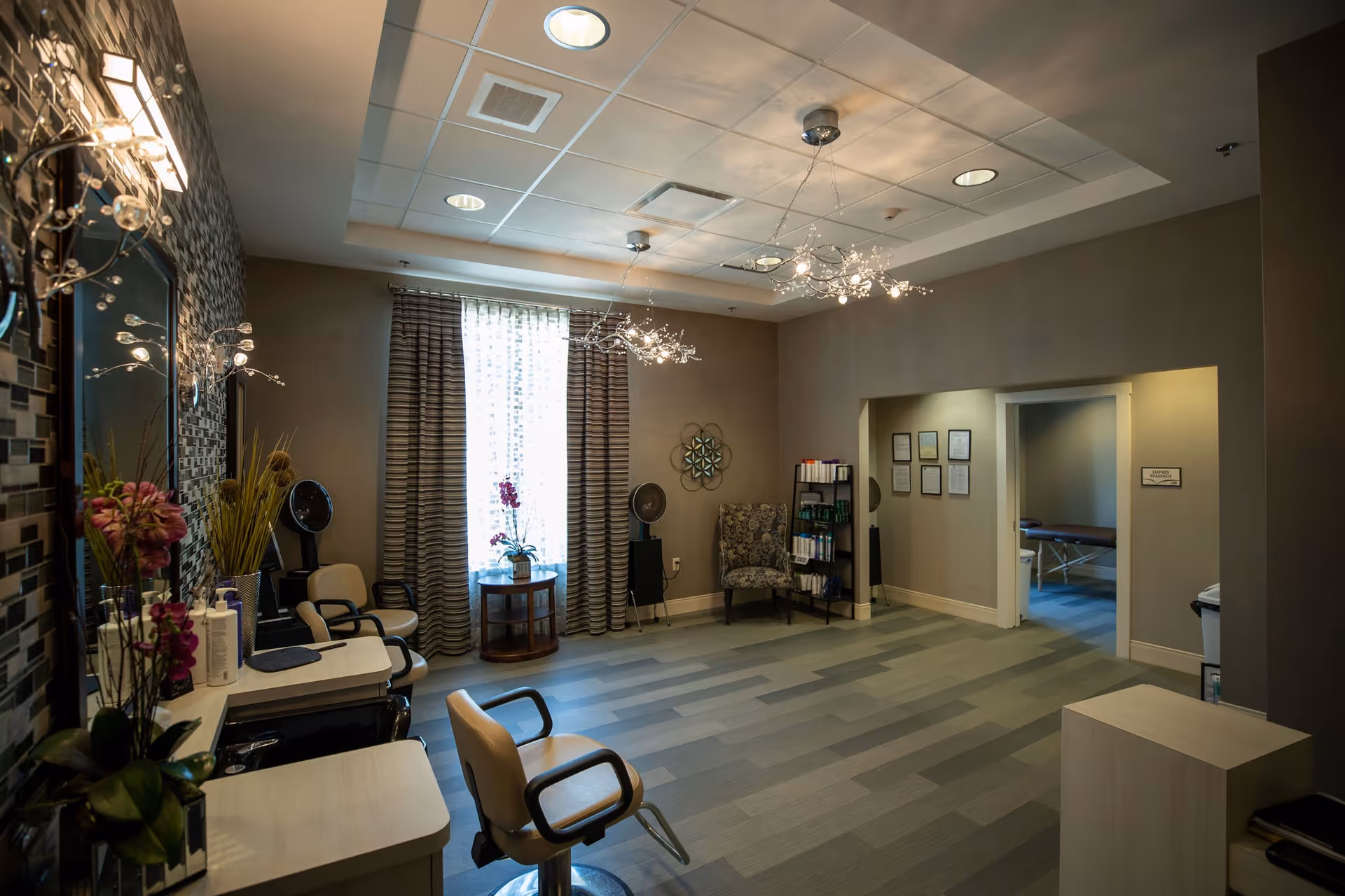 A well-lit salon or spa room with two salon chairs facing mirrors on the left side, decorative lighting fixtures on the ceiling, a window with striped curtains, a small round table with a flower pot, a patterned armchair, and a doorway leading to a massage room with a massage table visible.