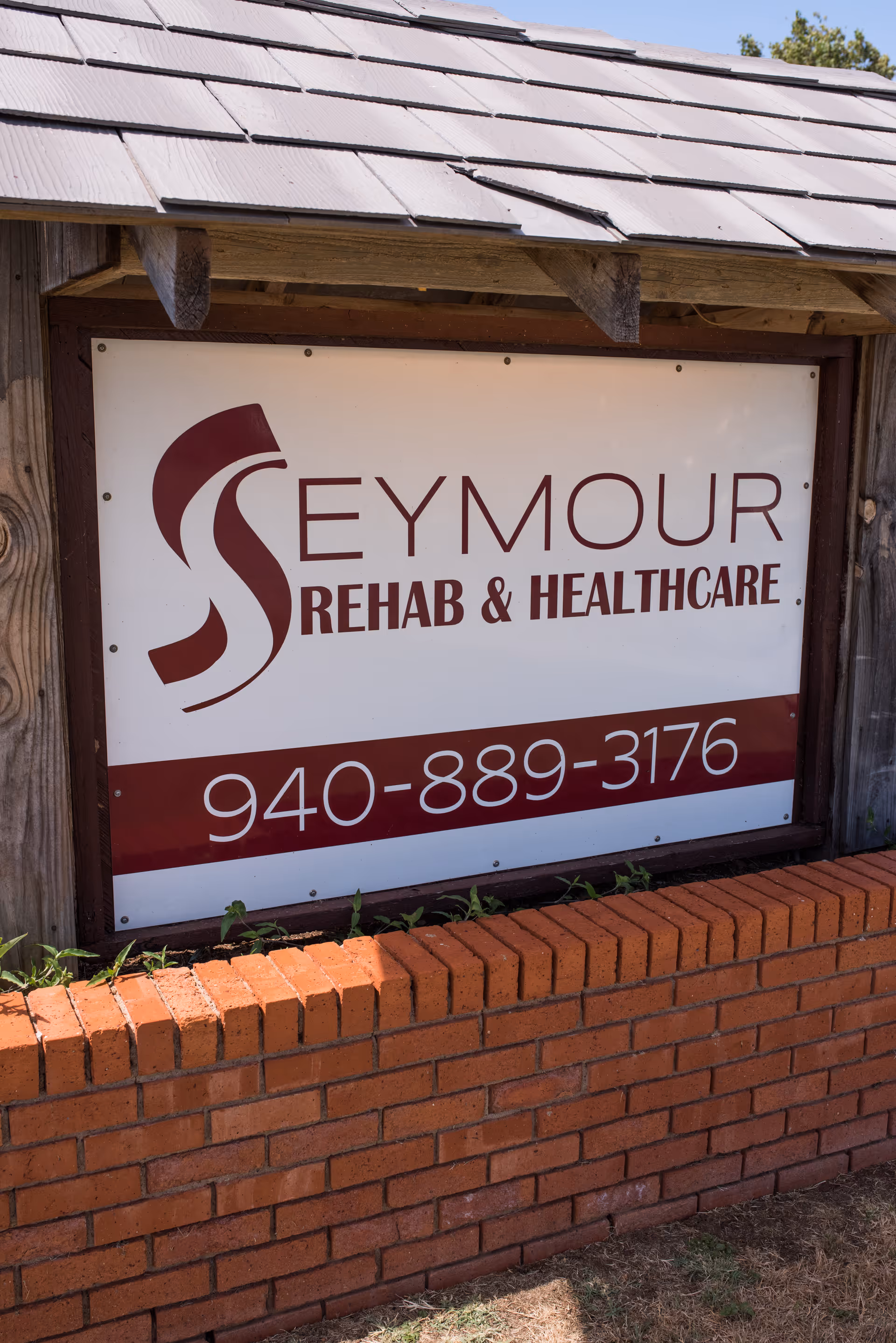 Outdoor sign for Seymour Rehab & Healthcare mounted on a wooden frame with a shingled roof above and a brick planter below, displaying the phone number 940-889-3176.
