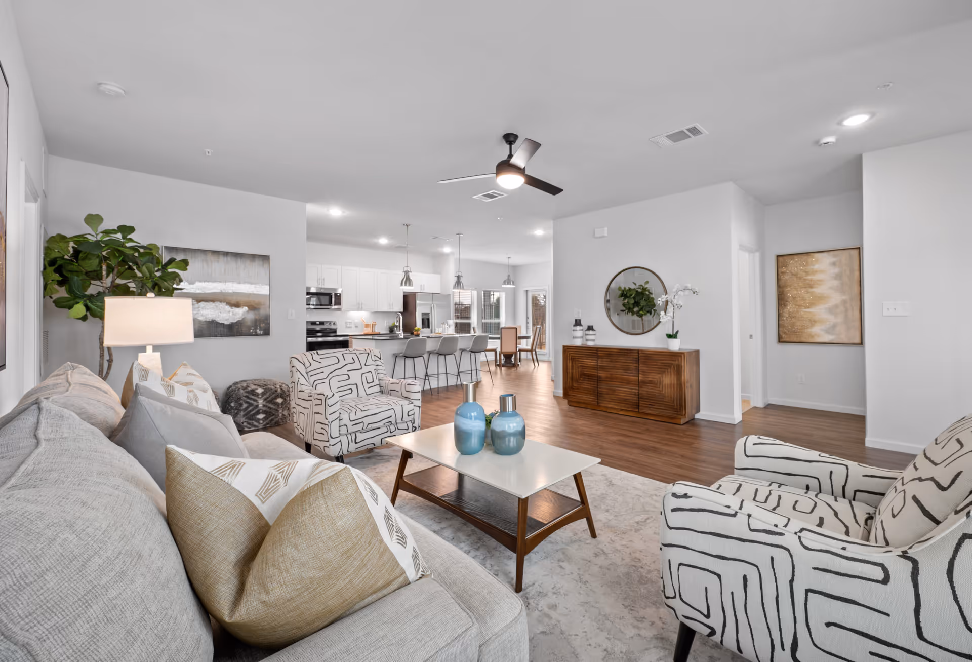 Open-plan living room with a sofa, patterned armchairs, coffee table and decor, looking toward a kitchen and dining area.