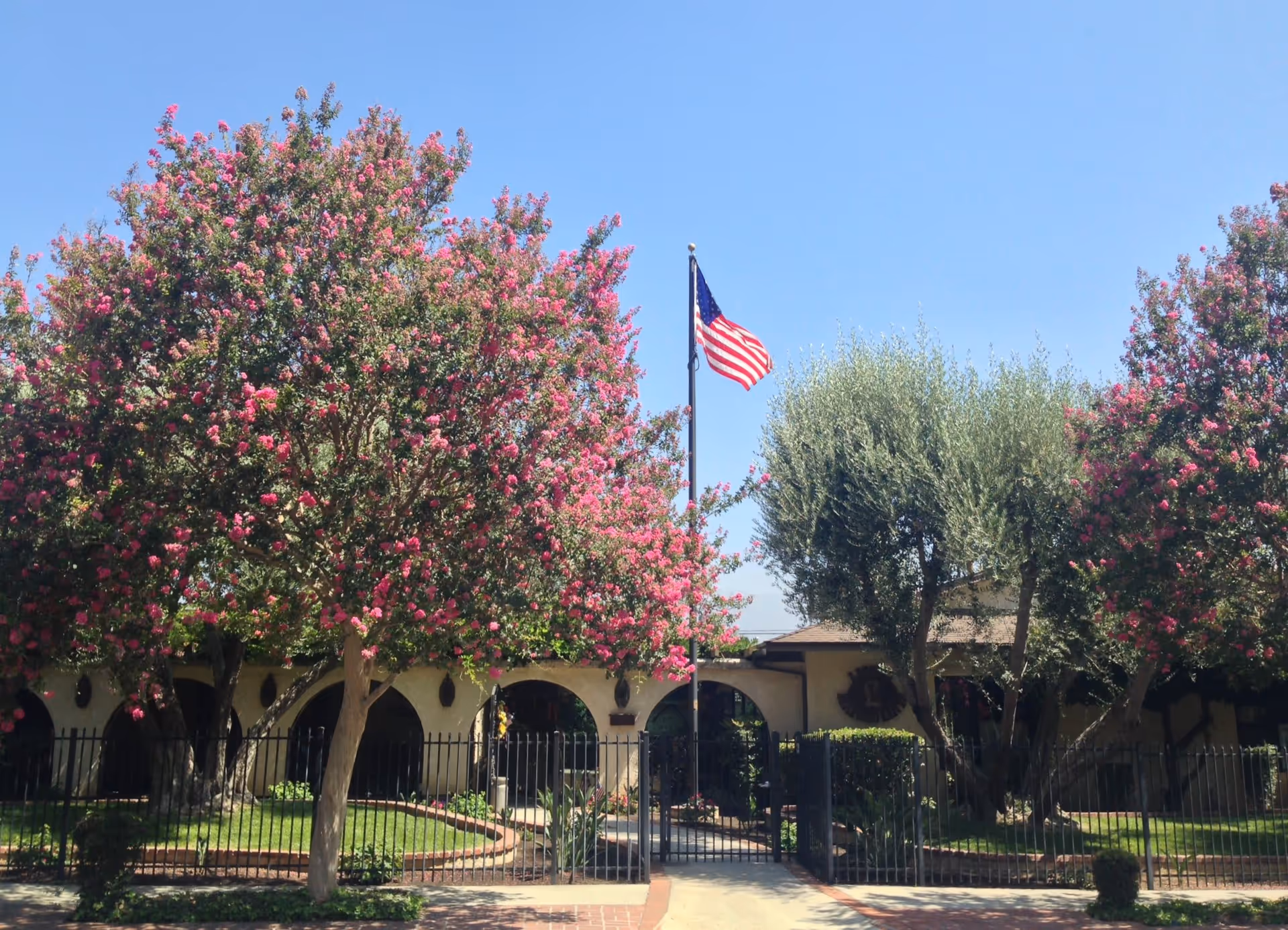 Front exterior view of a building with arched doorways, surrounded by a black metal fence and landscaped with green grass, pink flowering trees, and an American flag on a flagpole against a clear blue sky.