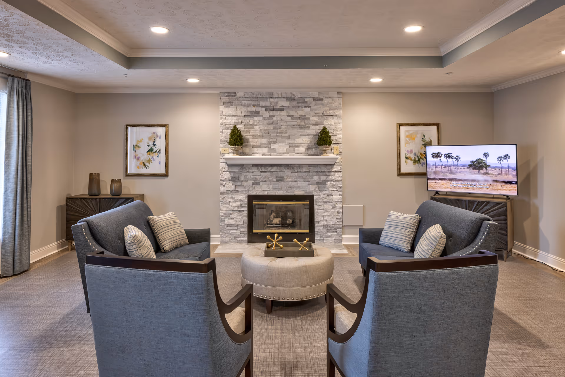 A cozy living room with two gray armchairs and two gray sofas arranged around a round beige ottoman. The room features a modern stone fireplace with two small decorative plants on the mantel. There are two framed floral artworks on the walls, a flat-screen TV on a stand showing a landscape with palm trees, and soft lighting from recessed ceiling lights. Gray curtains cover a window on the left side.