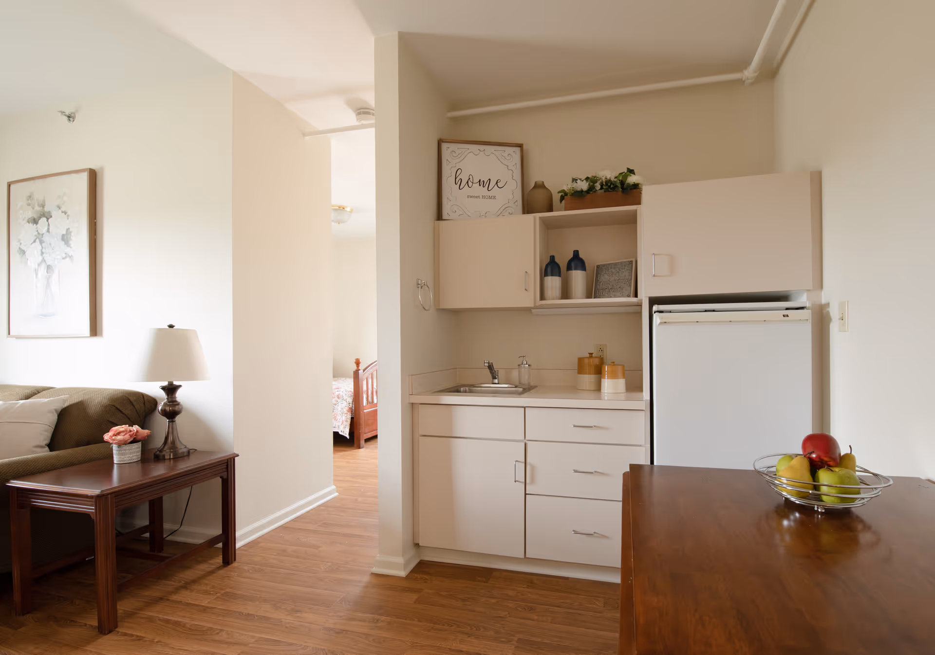A cozy interior space featuring a small kitchenette with white cabinets, a sink, and a mini refrigerator. On the countertop, there are decorative jars and a bowl of fruit. To the left, there is a wooden side table with a lamp and a small flower arrangement next to a green sofa. A framed floral picture hangs on the wall above the sofa. In the background, a bedroom with a wooden bed frame is partially visible through an open doorway.