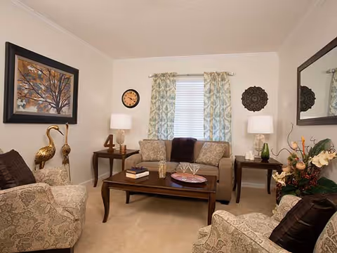 A cozy living room with a beige sofa and two patterned armchairs arranged around a wooden coffee table. The coffee table holds books, a decorative bowl, and a glass container. Two side tables with lamps flank the sofa. A window with patterned curtains is centered on the back wall, flanked by a clock and decorative wall art. A large mirror hangs on the right wall, and a floral arrangement is visible in the corner.
