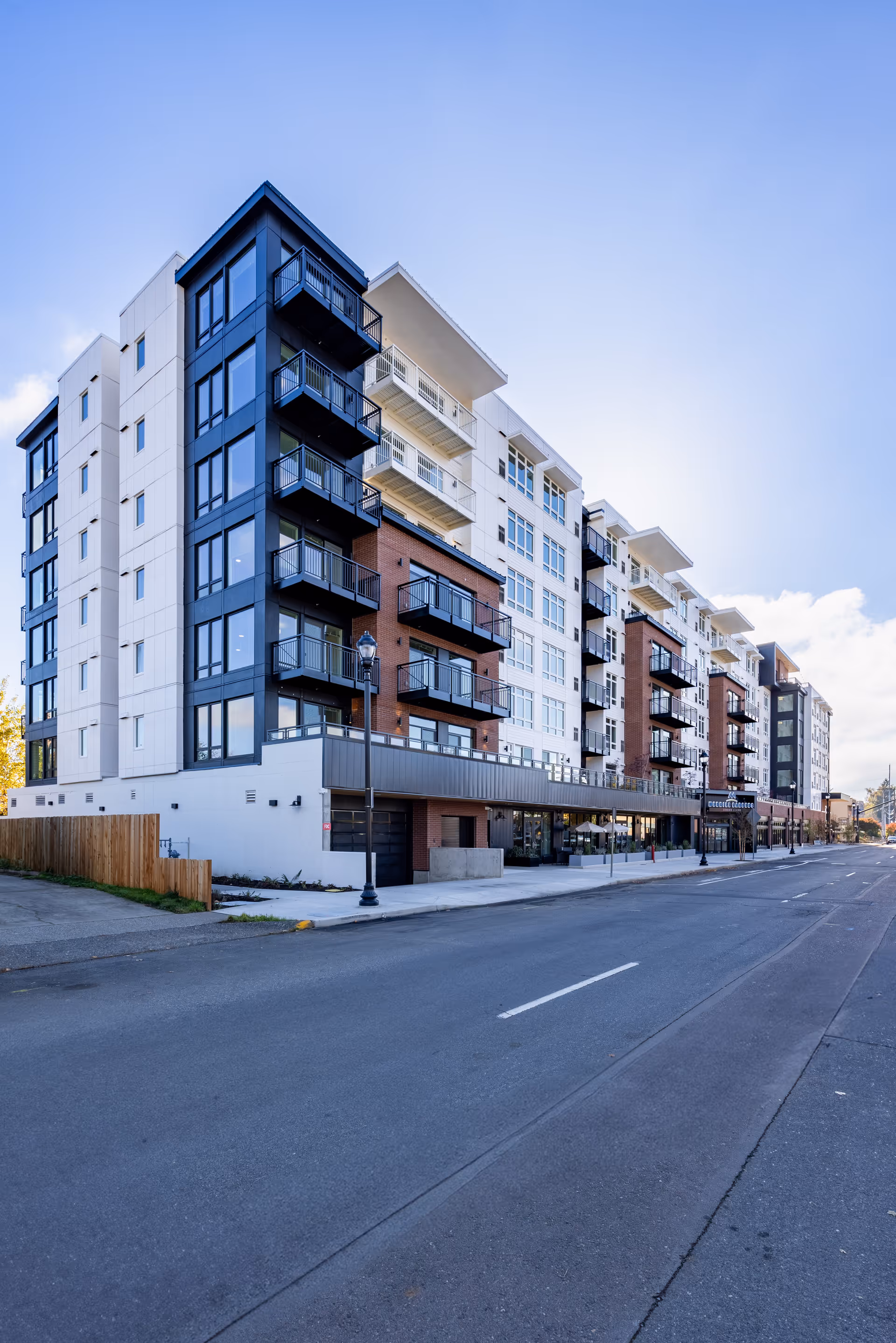 Exterior view of a modern multi-story residential building with balconies, large windows, and a mix of white, black, and brick facade under a clear blue sky.