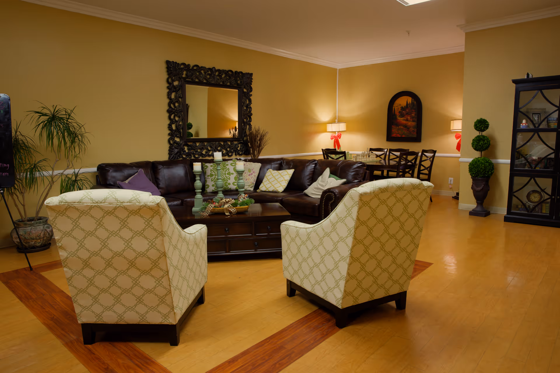 A cozy living room area with a dark brown leather sectional sofa adorned with various patterned pillows, two cream-colored armchairs with a geometric pattern, and a wooden coffee table with decorative candles and a centerpiece. The room has warm yellow walls, a large ornate mirror above the sofa, a dining table with chairs in the background, potted plants, and a black display cabinet with decorative items.