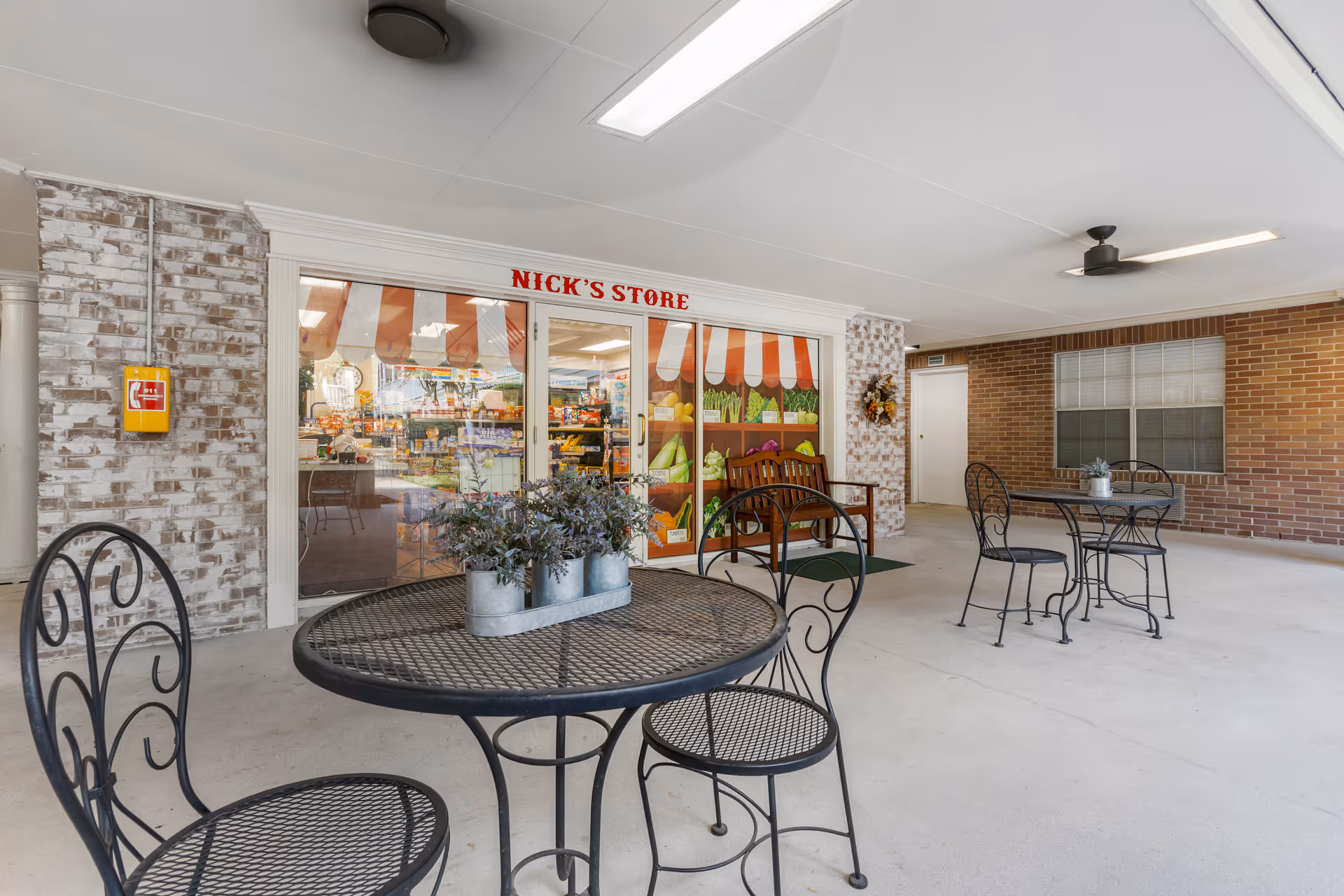 Covered outdoor seating area with black metal tables and chairs, potted plants on the tables, and a brick wall with a window and door. The door leads to Nick's Store, a small shop with a glass front and colorful awning design.
