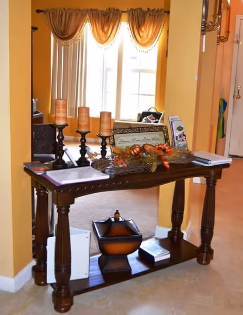A wooden console table with decorative items including three candle holders with candles, a framed sign, a fall-themed centerpiece, brochures, and books. The table is positioned in a warmly painted hallway with a window and curtains visible in the background.