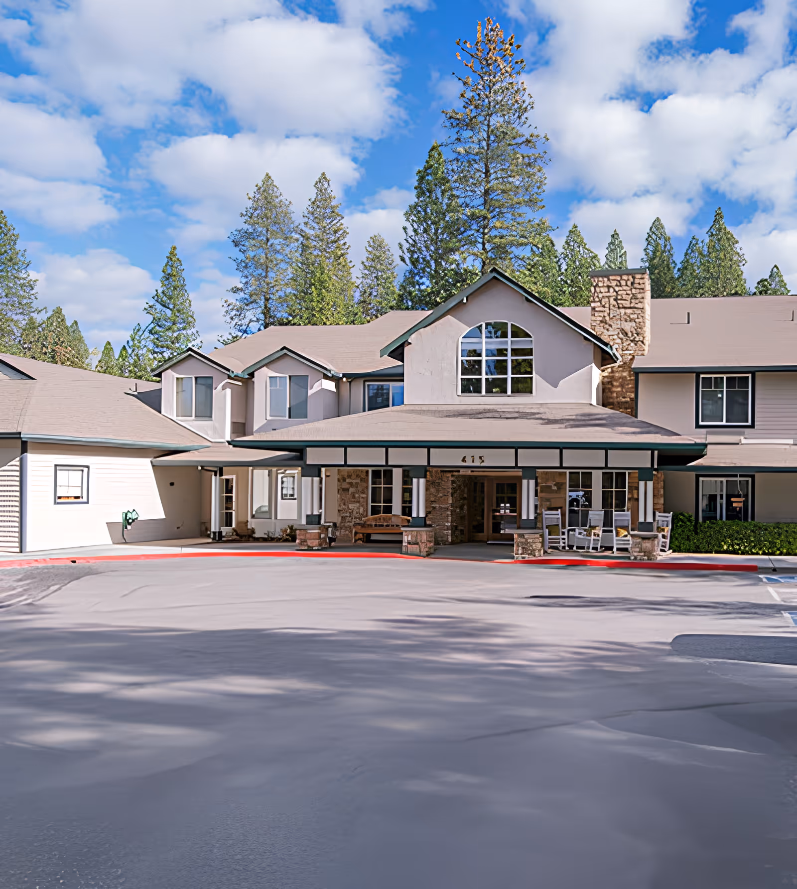 Front exterior view of Cascades of Grass Valley facility, showing a two-story building with a covered entrance, stone pillars, rocking chairs, and surrounded by tall pine trees under a partly cloudy blue sky.