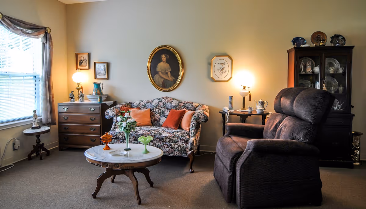 A cozy living room with a floral patterned sofa adorned with orange and red cushions, a dark brown recliner chair, a round marble-top coffee table with decorative items, a wooden chest of drawers with a lamp and figurines, and a glass-front cabinet displaying china. The walls are decorated with framed artwork and the room is softly lit by two table lamps.
