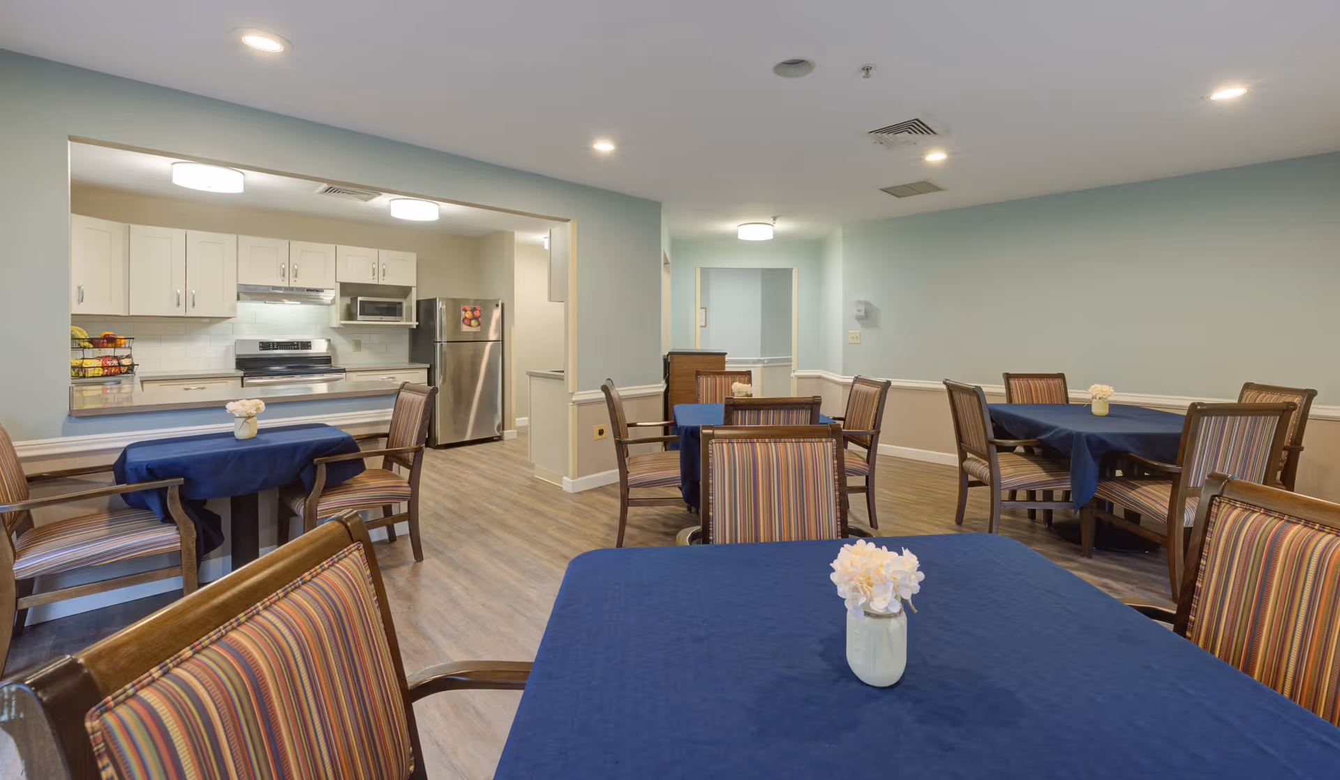 A dining area with several tables covered in blue tablecloths and wooden chairs with striped cushions. In the background, there is an open kitchen with white cabinets, a stainless steel refrigerator, stove, and microwave. The room has light blue walls and wood flooring.