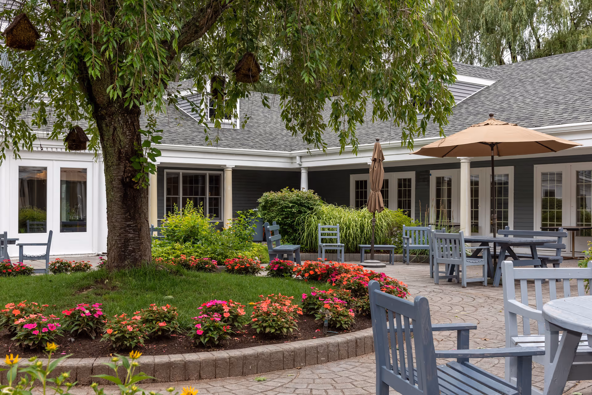 Outdoor patio area at Suffield House featuring a large tree with hanging birdhouses, colorful flower beds, gray wooden chairs and tables, and beige umbrellas. The building exterior is visible with multiple windows and white columns.