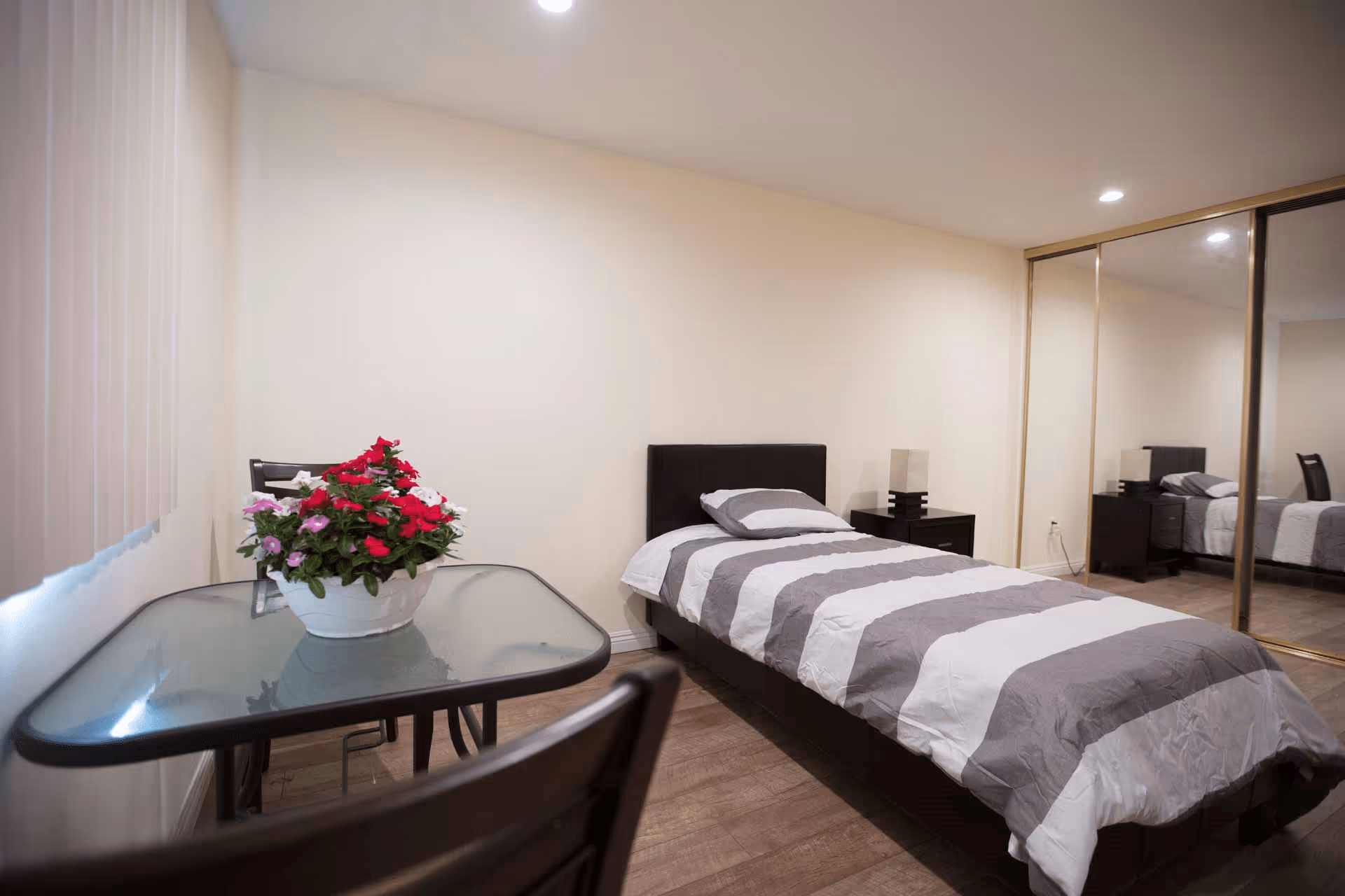 A simple bedroom with a single bed covered in a gray and white striped comforter, a matching pillow, a bedside table with a lamp, a glass-top table with a flower pot containing red, white, and pink flowers, wooden floor, and mirrored sliding closet doors.