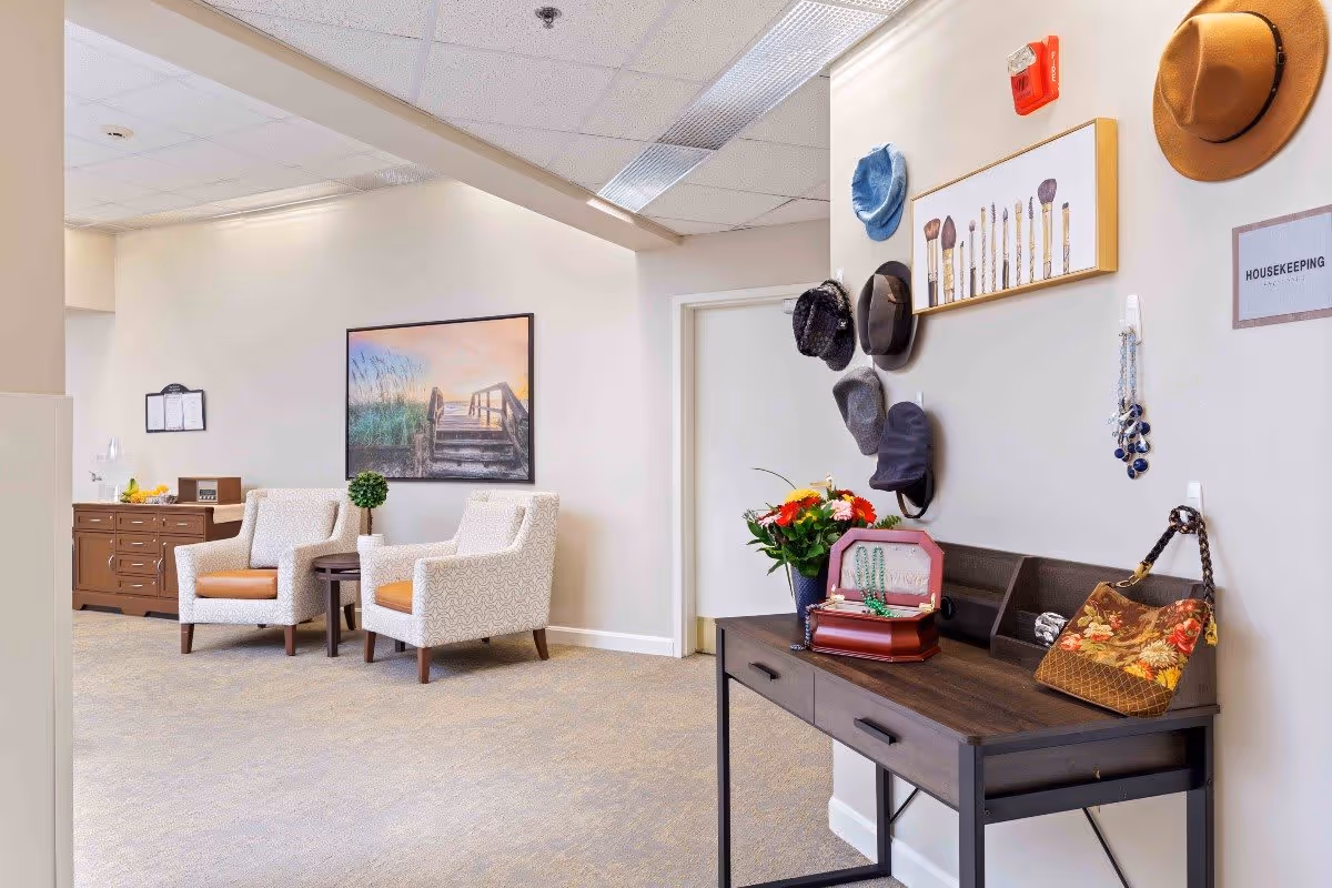 A cozy interior space with two patterned armchairs with brown cushions, a small round table with a potted plant between them, and a wooden sideboard with fruit and a lamp. On the right side, a dark wooden console table holds a flower vase, a jewelry box, and a floral handbag. Several hats and necklaces hang on the wall above the console table, along with a framed picture of makeup brushes and a sign labeled 'HOUSEKEEPING'. The walls are light-colored, and the floor is carpeted.