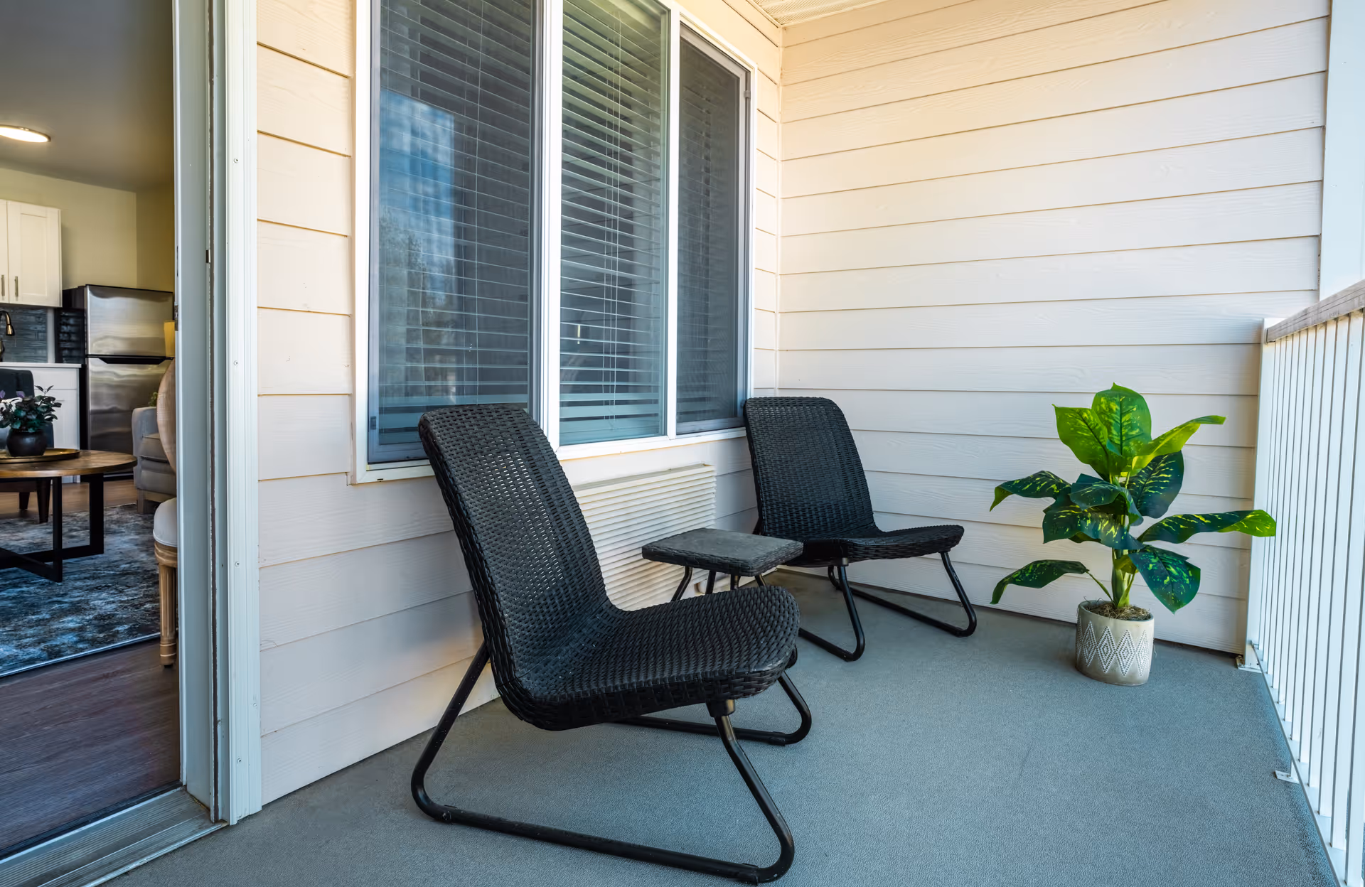 A small outdoor balcony area with two black woven chairs and a small matching table between them. There is a potted green plant on the right side near the railing. The balcony has light beige siding and a large window with closed blinds. The interior of a living room and kitchen is visible through an open door on the left.