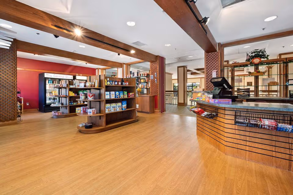 Interior view of a general store area inside a senior living facility with wooden shelves stocked with various grocery items and snacks. There is a checkout counter with a cash register and candy bars displayed on the front. The space has wooden flooring, exposed wooden beams on the ceiling, and a warm, inviting atmosphere.