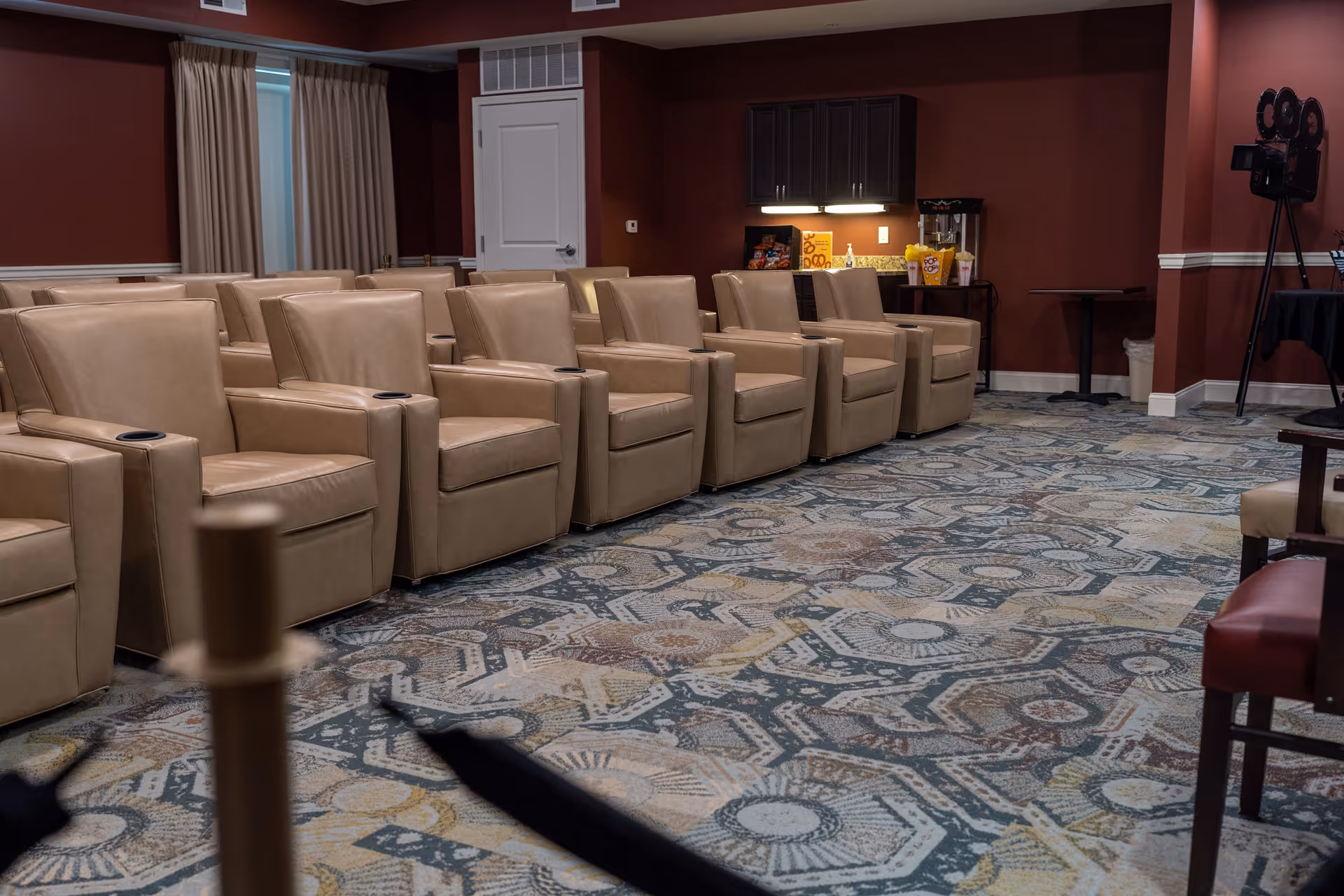 A room with multiple beige leather recliner chairs arranged in rows facing the same direction, a patterned carpet, dark red walls, a small counter with snacks and a popcorn machine, and a vintage film camera on a tripod in the corner.